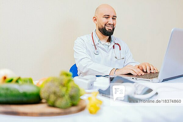 Lächelnde Ernährungsberaterin mit Laptop am Schreibtisch. Ernährungswissenschaftler Arzt mit Laptop am Arbeitsplatz. Bärtiger Ernährungswissenschaftler Arzt arbeitet am Laptop am Schreibtisch