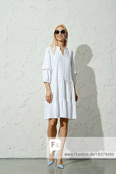 Positive Frau mit Sonnenbrille  die in einem weißen Baumwoll Sonnenkleid spazieren geht