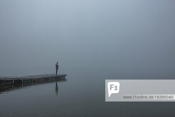 Blonde woman stands on dock of lake on foggy morning