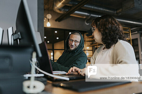 Junger männlicher Hacker diskutiert mit einer Kollegin am Schreibtisch in einem kreativen Arbeitsumfeld