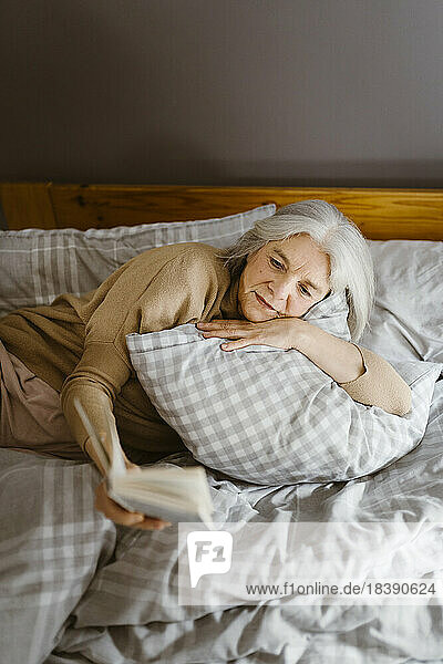 Ältere Frau liest ein Buch  während sie zu Hause auf dem Bett liegt