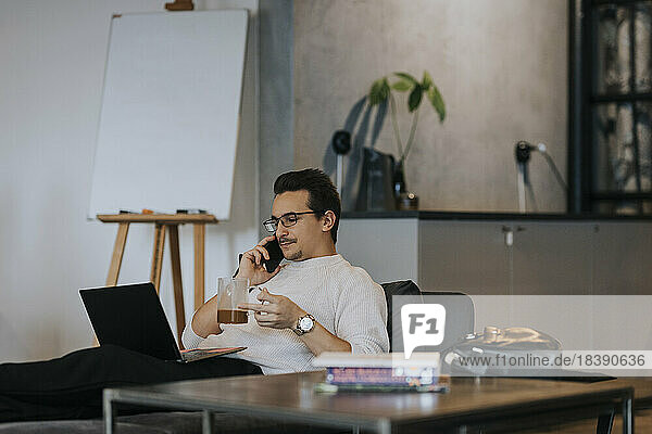 Geschäftsmann  der einen Kaffee trinkt  während er mit seinem Smartphone telefoniert und an seinem Laptop im Büro arbeitet