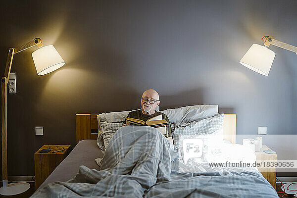 Senior man spending leisure time while reading book on bed at home