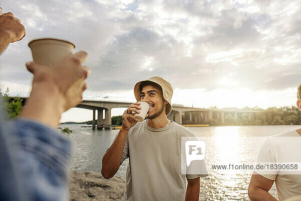Lächelnder junger Mann mit Eimerhut genießt ein Getränk mit einem Freund beim Picknick