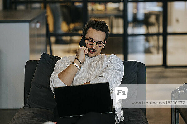 Junger Geschäftsmann  der mit seinem Handy telefoniert  während er an einem Laptop in einem kreativen Büro arbeitet
