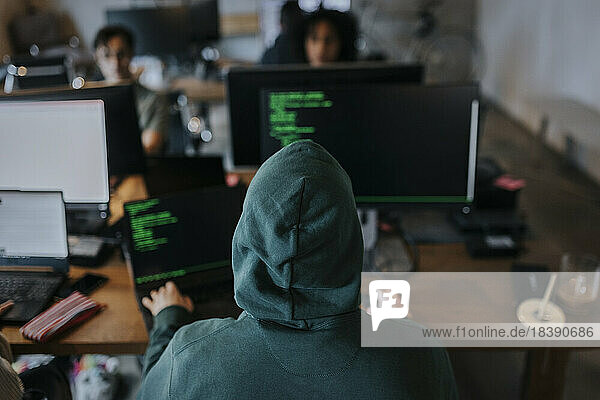 Rückansicht eines jungen männlichen Hackers mit Kapuze  der an einem Schreibtisch in einer kreativen Arbeitsumgebung am Computer programmiert