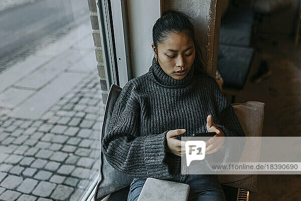 Junge Geschäftsfrau  die ein Mobiltelefon am Fenster im Büro benutzt