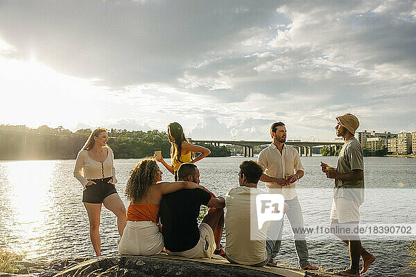 Multirassische männliche und weibliche Freunde unterhalten sich beim Picknick am Meer bei Sonnenuntergang