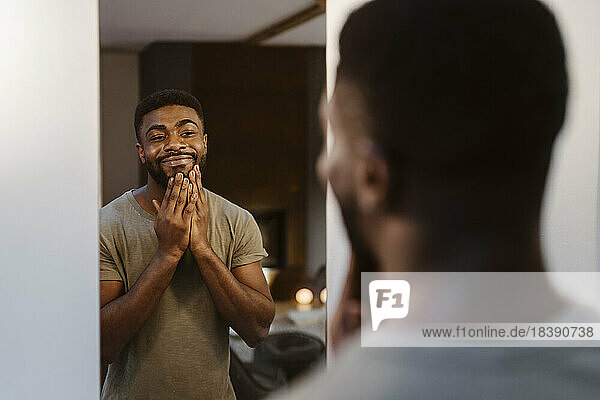 Lächelnder junger Mann  der sein Gesicht berührt  während er zu Hause in den Spiegel schaut