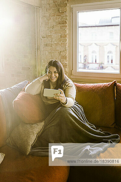Lächelnde Frau führt einen Videoanruf über ihr Smartphone durch  während sie zu Hause auf dem Sofa sitzt
