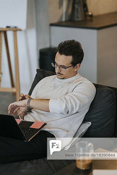 Junger Geschäftsmann  der die Zeit auf seiner Armbanduhr überprüft  während er mit seinem Laptop im Büro sitzt