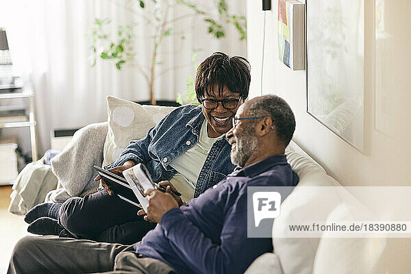 Happy senior woman with digital tablet talking to man sitting on sofa at home