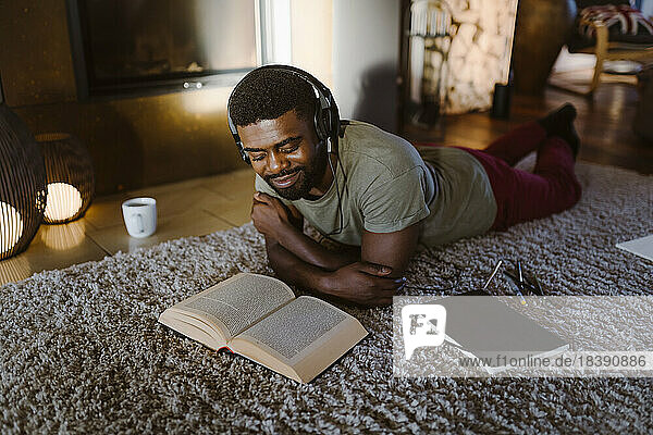 Smiling young man listening to music and reading book while lying on carpet at home