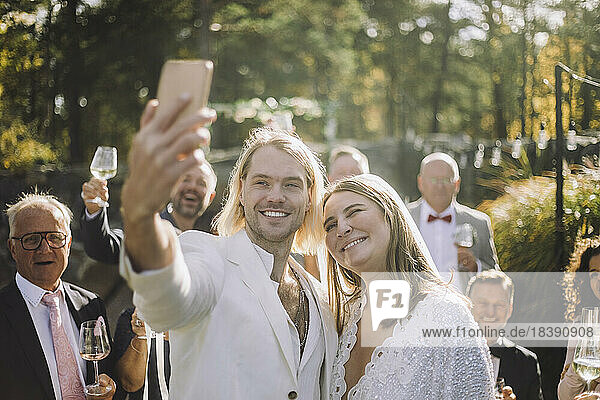 Glücklicher Bräutigam  der ein Selfie mit der Braut und den Gästen durch ein Smartphone bei der Hochzeitszeremonie macht