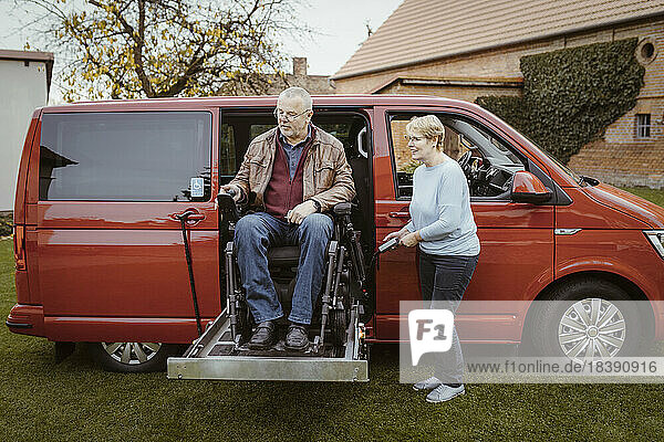 Lächelnde ältere Frau  die eine Fernbedienung in der Hand hält  während sie einem behinderten Mann im motorisierten Rollstuhl beim Aussteigen aus dem Auto hilft.