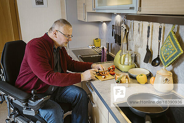 Älterer Mann im Ruhestand mit Behinderung sitzt im motorisierten Rollstuhl und schneidet Paprika am Küchentisch