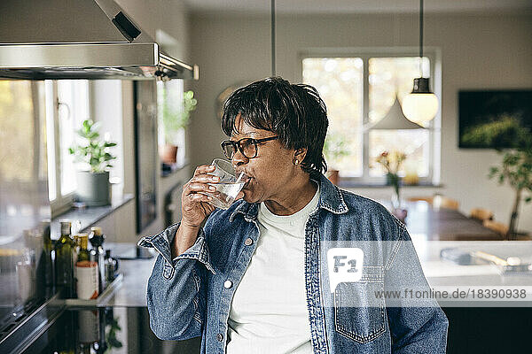 Ältere Frau in Jeansjacke trinkt Wasser in der Küche zu Hause