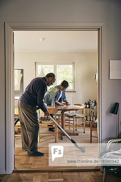 Seitenansicht eines älteren Mannes  der den Boden mit einem Staubsauger reinigt  gesehen durch eine Türöffnung zu Hause