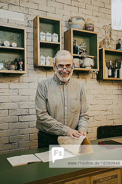 Porträt eines lächelnden älteren männlichen Besitzers  der eine Schachtel in einem Geschäft für Wohnkultur einpackt