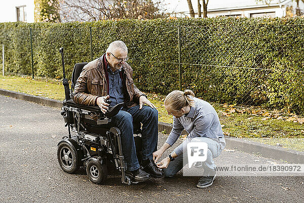 Junger Mann kniet beim Binden der Schnürsenkel seines behinderten Vaters  der im motorisierten Rollstuhl sitzt