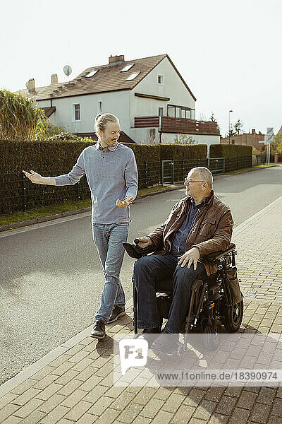 Lächelnder älterer Mann mit Behinderung in einem motorisierten Rollstuhl  der von einer jungen Betreuerin bei Sonnenschein auf der Straße geführt wird  in voller Länge