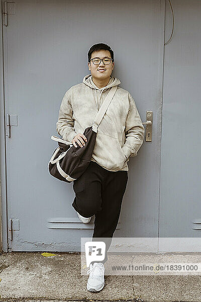 Lächelnder junger Mann mit Hand in der Tasche  der sich an eine graue Tür lehnt