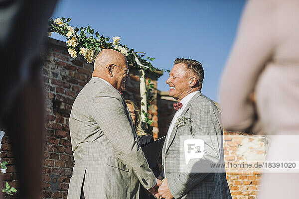 Glückliches frisch verheiratetes homosexuelles Paar  das sich bei der Hochzeit an den Händen hält  während eines sonnigen Tages