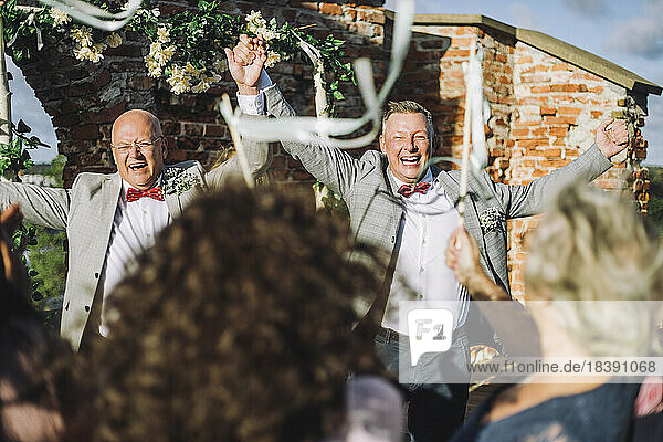 Fröhliches frisch verheiratetes schwules Paar  das sich an den Händen hält  während es seine Hochzeitszeremonie mit den Gästen genießt