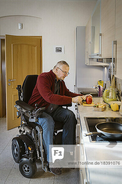 Rentner mit Behinderung sitzt im motorisierten Rollstuhl und schneidet Paprika an der Küchentheke in voller Länge