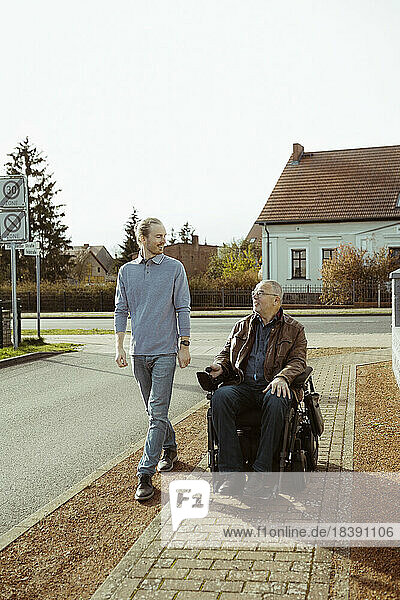 Lächelnder älterer Mann mit Behinderung im motorisierten Rollstuhl  der von einer jungen Betreuerin auf der Straße bei sonnigem Wetter begleitet wird