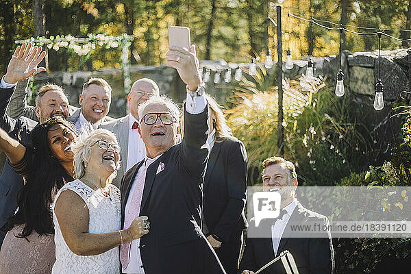 Happy senior man taking selfie with bride and family on smart phone during wedding ceremony