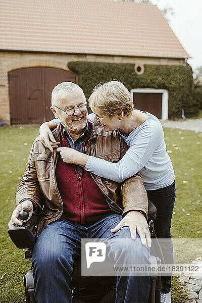 Fröhliche ältere Frau im Ruhestand umarmt Mann mit Behinderung  der im motorisierten Rollstuhl im Hinterhof sitzt