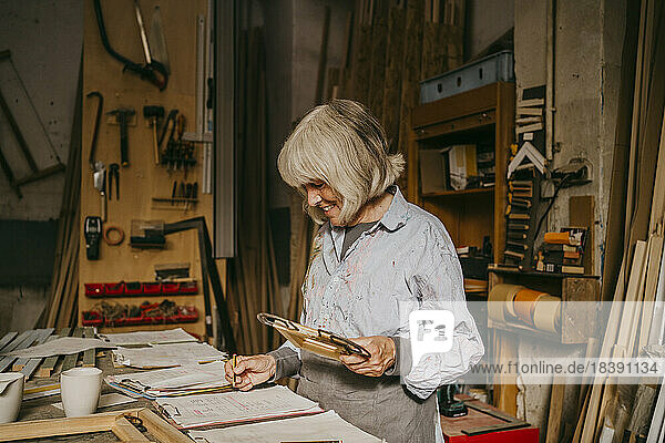 Lächelnder älterer weiblicher Schreiner  der einen Rahmen hält  während er in der Werkstatt auf einem Klemmbrett schreibt