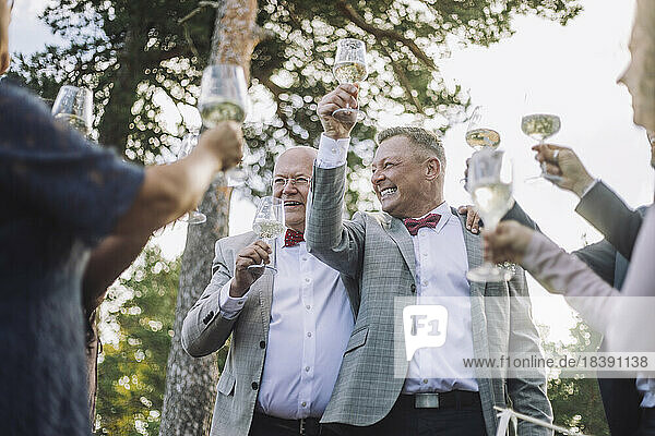 Glückliches frisch verheiratetes schwules Paar erhebt seine Weingläser mit Familie und Freunden bei der Hochzeit