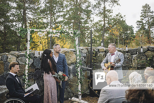 Älterer Mann spielt Gitarre für frisch vermähltes Paar von Gästen und Minister während der Hochzeitszeremonie