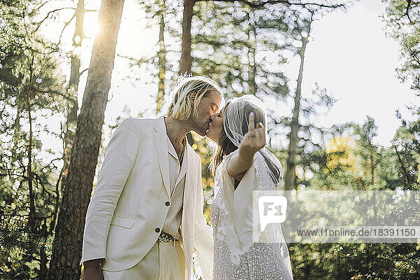 Braut zeigt Ringfinger und küsst Bräutigam auf den Mund im Wald während der Hochzeit