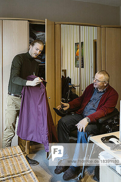 Junge männliche Pflegekraft zeigt einem älteren Mann mit Behinderung  der zu Hause im motorisierten Rollstuhl sitzt  ein Hemd