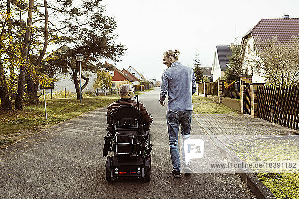 Rear view of senior man with disability in motorized wheelchair by young caregiver walking on road