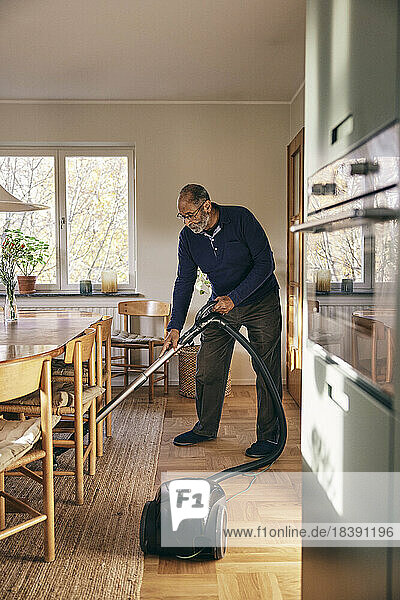 Älterer Mann reinigt Teppich mit Staubsauger im Stehen zu Hause
