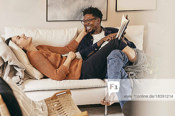 Happy multiracial couple having fun while reading books on sofa at home