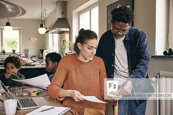 Multiracial couple discussing over financial bills while sons in background at home