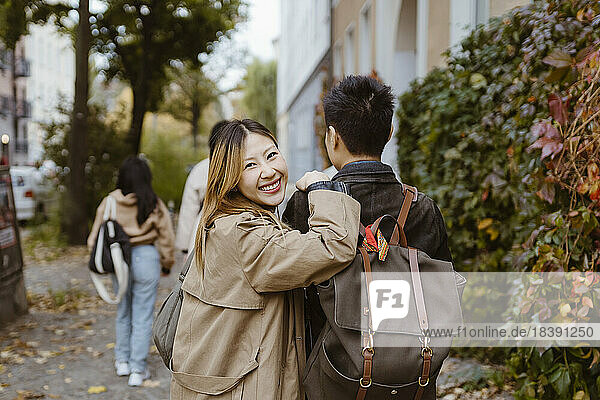 Porträt einer glücklichen Frau  die über die Schulter schaut  während sie mit einem männlichen Freund auf dem Gehweg spazieren geht