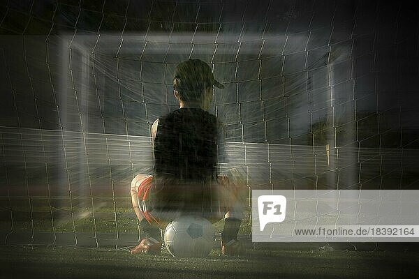 Kleiner Junge von hinten  im Fußballtrikot  auf Fußball sitzend  im Hintergrund ein Tor und das Netz  geisterhaft  verschwommen