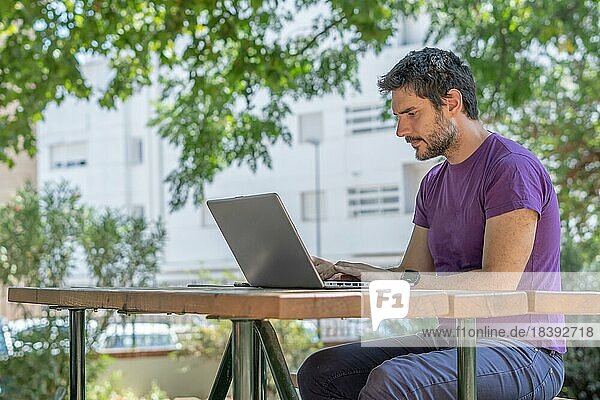 Young man with beard  in profile  working with his pc at a wooden table in the park
