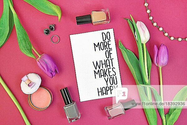 Happiness motivierendes Konzept mit Zeichen sagen Tun Sie mehr von dem  was Sie glücklich macht  umgeben von Frühlingsblumen und weibliche Accessoires wie Makeup auf rosa Hintergrund  flach legen