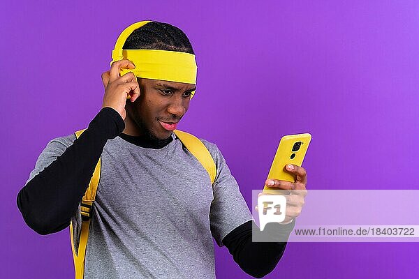 Schwarz ethnischen Mann mit Rucksack und gelbe Kopfhörer auf einem lila Hintergrund  Student Konzept  mit dem Telefon ernst