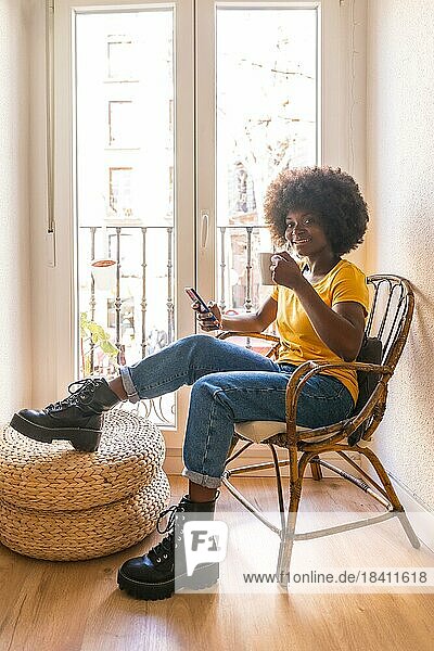 Afrohaarige Frau am Fenster zu Hause mit einem heißen Getränk  Kaffee am Morgen mit Blick auf das Handy