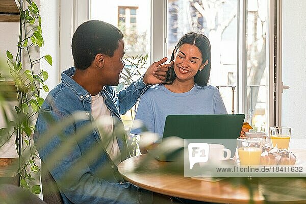 Verliebtes multiethnisches Paar  das beim Frühstück am Fenster einen Online Einkauf über den Computer tätigt