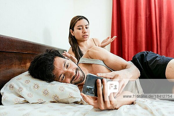 Husband with cell phone ignoring wife in bed. Annoyed woman with husband while holding cell phone in bed. Cheating Husband Texting On Phone Ignoring Wife Lying In Bed