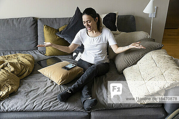 Frau mit Behinderung gestikuliert  während sie auf dem Sofa sitzend ihr Smartphone betrachtet